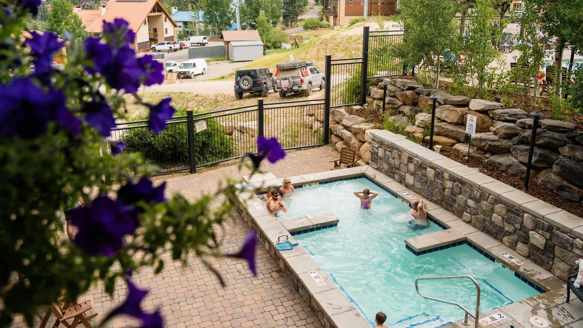 A lively swimming pool scene, with people enjoying the water and colorful flowers in the foreground, all set amid lush natural surroundings. The atmosphere is joyful and sociable, encouraging relaxation and group fun.