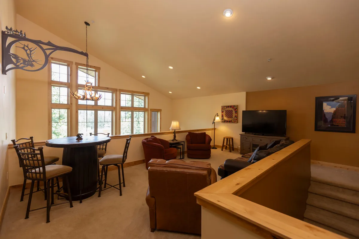 A spacious interior lounge featuring large windows that allow natural light to pour in, a comfortable seating area with a sofa facing a TV setup, and a bar table with chairs nearby. The arrangement suggests flexibility for both entertainment and relaxation, with space for informal gatherings or cozy downtime. The atmosphere is inviting, with relaxed, family-friendly energy.