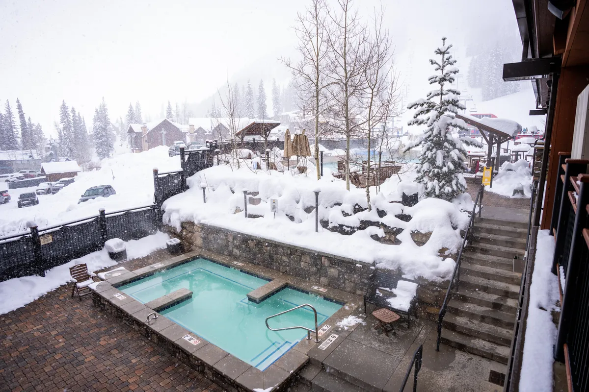 A steaming hot tub set in a snowy landscape with pine trees and inviting outdoor seating. The snow-covered surroundings evoke après-ski relaxation in a quintessential mountain setting.
