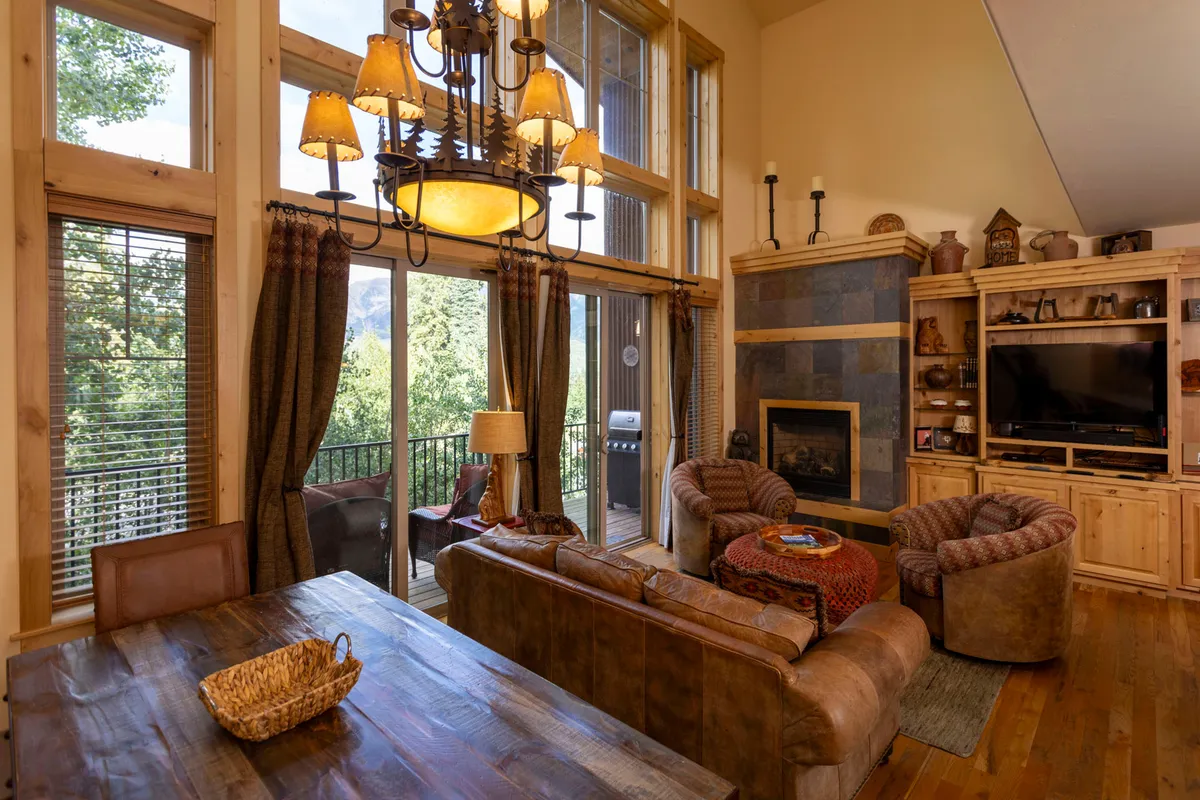 A spacious, open-concept living area with large windows offering panoramic mountain views. The room features a comfortable seating area centered around a stone fireplace, a stylish chandelier, a generous dining table, and a seamless connection between the living, dining, and lounging spaces. Natural light pours in, creating an inviting atmosphere perfect for gatherings.