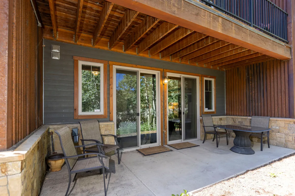 An inviting exterior scene showing a stone-walled patio just outside sliding glass doors, with a cozy outdoor seating area perfect for relaxing while listening to the creek. The area is surrounded by trees and bathed in tranquil morning light.