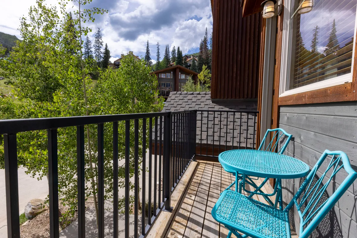 This exterior image captures a turquoise bistro table and chairs set on a balcony, framed by lush green trees and a stunning mountain backdrop. The scene suggests an inviting space to relax outdoors, take in the views, and enjoy the fresh alpine air.